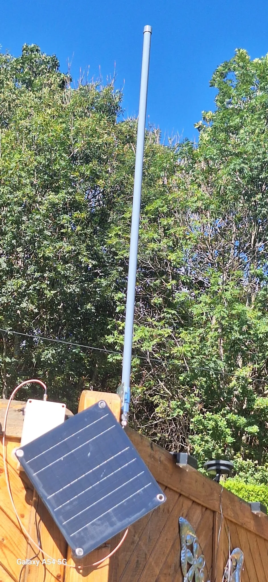 Fiberglasantenne an einem Holzgeländer, daneben weißes Gehäuse und schräg montiertes Solarpanel