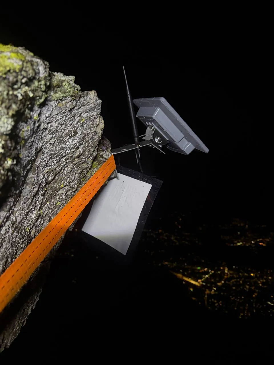 Solar-Node bei Nacht mit orangem Spanngurt an einem Felsen am Martennock befestigt, darunter die Lichter des Drautals