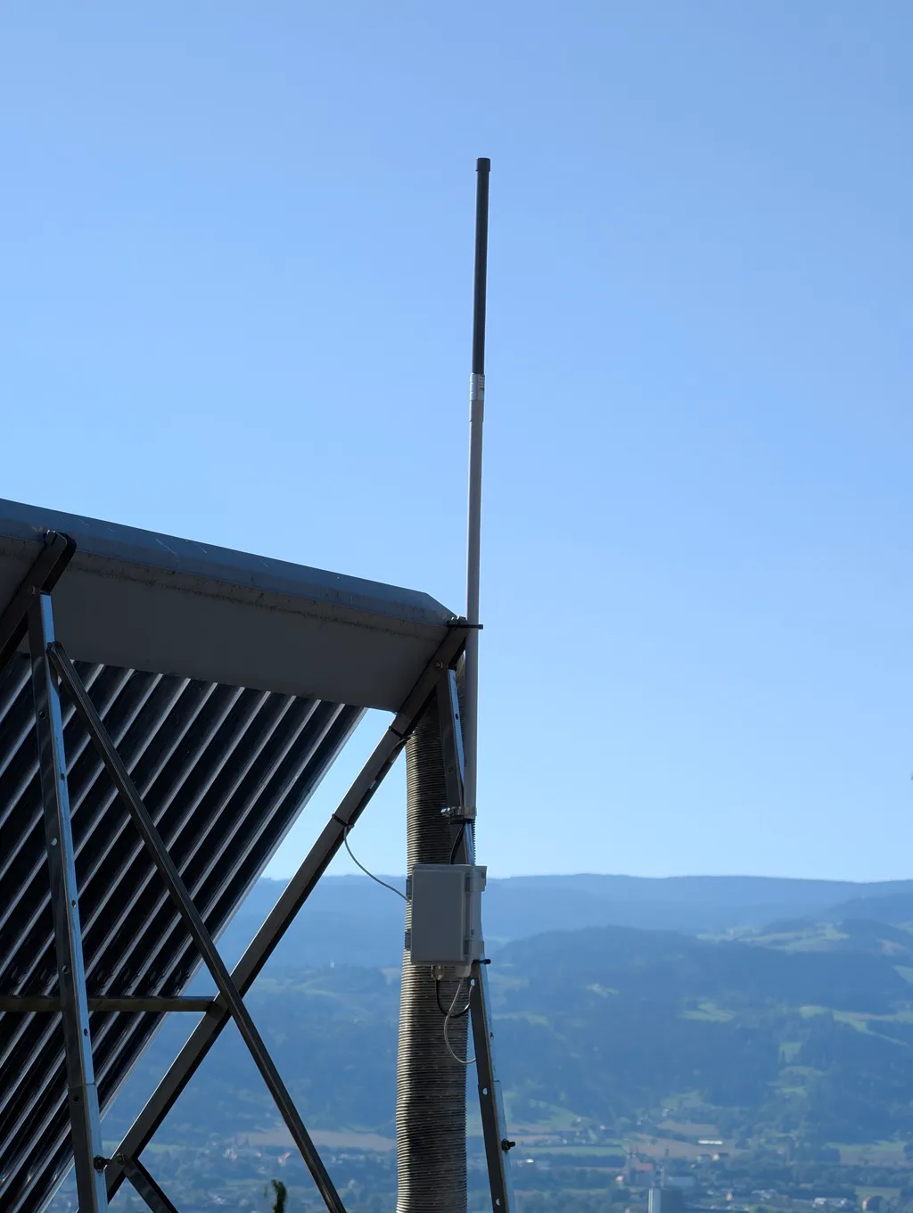 Fiberglasantenne und Gehäuse an einem Mast neben Solarkollektoren, dahinter Talblick